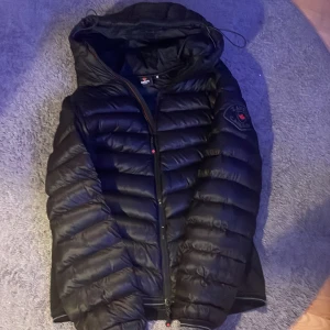 Svart pufferjacka från Zavetti Canada - Säljer en svart pufferjacka från Zavetti Canada i storlek M. Jackan har huva, breda quiltade sektioner och en snygg logga med röd lönnlöv på ärmen. Dragkedja framtill och två sidofickor. Perfekt för kalla dagar och riktigt stilren look.