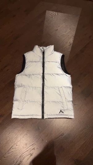 Vit dunväst från McKinley 146/152 - Snygg vit dunväst från McKinley med svart dragkedja och svarta detaljer vid ärmöppningarna. Västen har två fickor framtill och en stilren logga nere till höger. Perfekt för lager-på-lager under kyliga dagar.
