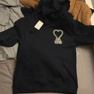 Mörkblå hoodie från Ami Paris XL - Snygg mörkblå hoodie från Ami Paris i storlek XL med ikonisk hjärta-A patch på bröstet. Tröjan har huva med snörning och en stor magficka. Materialet är mjuk bomull och passformen är normal. Perfekt för dig som gillar stilrena streetwear-plagg.