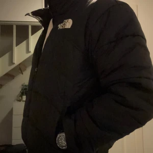 Svart pufferjacka från The North Face - Säljer en svart pufferjacka från The North Face med klassisk logga på bröstet och ryggen. Jackan har hög krage, dragkedja framtill och två sidofickor med dragkedja. Perfekt för kalla dagar och har en clean, stilren look som funkar till allt.