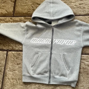 Grå Magnifique hoodie med dragkedja - Snygg grå hoodie från Magnifique med vit text på bröstet. Tröjan har huva, praktiska fickor framtill och hel dragkedja. Perfekt för chill dagar och streetstyle. Mjuk bomullskänsla och klassisk passform.