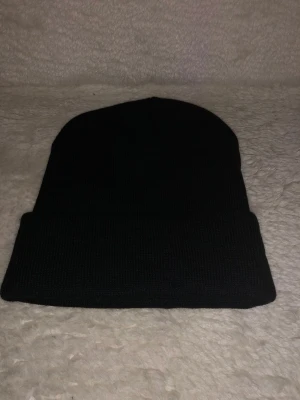 Svart stickad mössa från Lacoste - Svart mössa från Lacoste med klassisk uppvikt kant och den ikoniska gröna krokodilloggan framtill. Stickad i mjukt material som håller dig varm och snygg under kyliga dagar. Perfekt för dig som gillar stilrena accessoarer med en sportig touch.