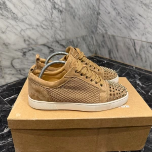 Christian Louboutin | Unika beige - Säljer ett par beiga sneakers från Christian Louboutin i mocka med ikoniska röda sulor och coola nitar på tån. Skorna har snörning och vit platt sula, perfekt för dig som vill sticka ut med lyxig streetstyle. Grym färg och detaljer som verkligen syns. Dustbag medföljer, låda, och allt annat, möts på Ostermalm eller fraktar