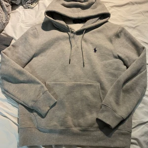 Grå hoodie från Polo Ralph Lauren - Säljer en grå hoodie från Polo Ralph Lauren med lila logga broderad på bröstet. Tröjan har huva med snörning, stor magficka och ribbade muddar vid ärmslut och nederkant. Perfekt för chill dagar och enkel att matcha med jeans eller joggers.
