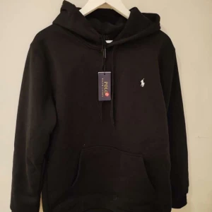 Svart hoodie från Polo Ralph Lauren - Svart hoodie från Polo Ralph Lauren med klassisk huva och snörning. Tröjan har en vit broderad logga på bröstet och en stor magficka framtill. Perfekt för dig som gillar stilrena och bekväma plagg med streetkänsla.