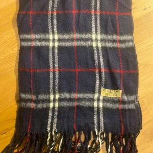 Burberry halsduk - Snygg halsduk från Burberry i 100% kashmir, med klassiskt rutigt mönster i marinblått, vitt och rött. Mjukt material och fransar i ändarna. Perfekt för att ge din outfit en lyxig touch och hålla dig varm under kyliga dagar.