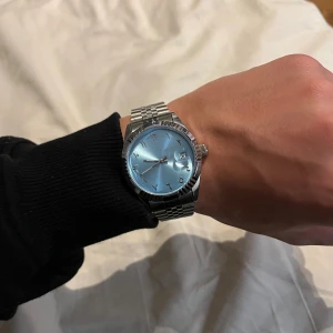 Stilren klocka med blå urtavla - Säljer en snygg klocka med ljusblå urtavla och datumvisning. Klockan har ett klassiskt runt silverfärgat boett och länkarmband i metall. Indexen är markerade med stilrena siffror och armbandet har en robust låsning. Perfekt accessoar för dig som gillar stilren design.