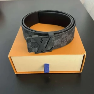Louis Vuitton rutig svart bälte - Snyggt Louis Vuitton bälte med klassiskt rutigt mönster i mörkgrå och svart. Bältet har en stilren LV-logga i matt svart metall som spänne. Materialet är skinn och designen är både modern och tidlös, perfekt för dig som gillar exklusiva accessoarer.