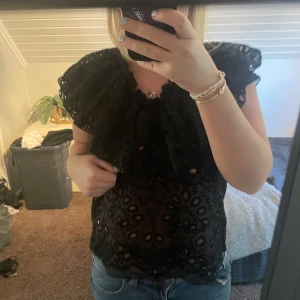 Svart blus med volang och spets - Säljer en svart blus med stora volangdetaljer vid axlarna och genombruten spets med mönster. Från Carin Wester