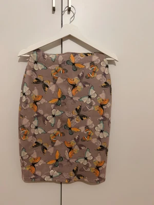 Kort kjol med fjärilsmönster från Chicing - Säljer en snygg kort kjol från Chicing med ett färgglatt fjärilsmönster i orange, turkos, svart och beige på en ljuslila bakgrund. Kjolen har en rak passform och är tillverkad i ett mjukt syntetmaterial som känns skönt mot huden. Perfekt för dig som gillar unika prints!