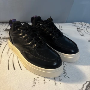 Svarta sneakers från Eytys Siba - Snygga svarta Eytys Siba sneakers med chunky vit sula och lila hälflik. Ovandel i skinn och mesh, klassisk snörning och perforerade detaljer på tån. Perfekt för dig som gillar streetstyle och vill ha en cool och modern look.