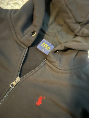 Svart fleece från Polo Ralph Lauren - Svart fleece från Polo Ralph Lauren i storlek XL med klassisk röd logobrodyr på bröstet. Tröjan har huva med snörningshål, hel dragkedja framtill och två stora fickor. Materialet är mjuk bomull, perfekt för kallare dagar. 