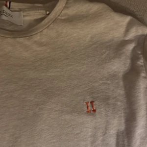 Vit t-shirt från Les Deux - Snygg vit t-shirt från Les Deux med klassisk rund hals och korta ärmar. På bröstet finns en liten broderad röd II-logga som ger en clean och stilren look. Tillverkad i mjuk bomull som känns skön mot huden. Perfekt för dig som gillar minimalistisk stil.