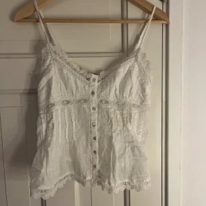 Vit linnetopp med spets och knappar - Säljer denna supersöta vita linnetopp med tunna axelband och spetsdetaljer runt om, med knappar fram till. Säljer då den inte kommer till användning💓