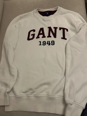 Vit sweatshirt från GANT med tryck - Vit sweatshirt från GANT med stort vinrött GANT-tryck och blått årtal 1949 på bröstet. Tröjan har rund hals, ribbade muddar och är långärmad. Perfekt för dig som gillar klassisk och sportig stil.