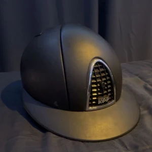 Svart ridhjälm från KEP - Snygg svart ridhjälm från KEP med matt finish och ventilerad front i metall-look. Klassisk rundad form med bred skärm och justerbart hakband. Perfekt för dig som vill ha både stil och säkerhet i stallet. Skitig på bild men rengörs innan försäljningen.