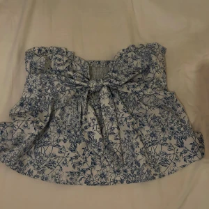 Blommig vit och blå tubtopp med volang - Supersöt tubtopp i vitt med blått blommönster och volangkant upptill. Toppen har en stor knytning framtill och smockad rygg för skön passform. Materialet känns lätt och luftigt, perfekt för varma dagar. Passar dig som gillar romantisk och feminin stil.