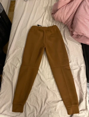 Nike Tech Fleece set i brun - Nike Tech Fleece set i snygg brun färg. Hoodien har dragkedja, huva och svarta detaljer vid fickorna. Byxorna har smal passform, svarta dragkedjor på fickorna och snörning i midjan. Byxorna är i storlek S och Hoodien är i storlek M