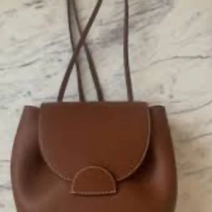 Brun axelväska från Polène i skinn - Snygg brun axelväska från Polène i äkta skinn med rundad form och lock med dekorativ söm. Väskan har justerbar axelrem och guldfärgade detaljer. Perfekt storlek för det viktigaste och stilren design som lyfter din outfit. With box and dustbag