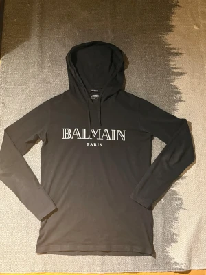 Balmain hoodie - Svart hoodie från Balmain Paris med stort vitt logotryck på bröstet. Tröjan har huva, långa ärmar och är tillverkad i mjuk bomull. Skick 9,5/10 Inpricnip i nyskick och använd ett fåtal gånger.