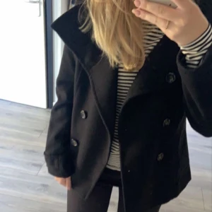 Svart skepparkavaj från H&M, strl 38 - Svart skepparkavaj från H&M i storlek 38. Jackan har bred krage, dubbelknäppning med stora svarta knappar och långa ärmar. Materialet är ullblandning som ger en klassisk och stilren look. Perfekt för dig som gillar tidlös stil och vill ha en jacka med snygg siluett.