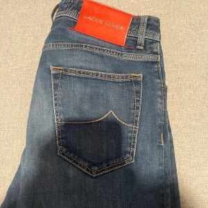 Jacob Cohën blå jeans, stl 33 - Snygga blå jeans från Jacob Cohën i klassisk femficksmodell. Jeansen har en mörkblå tvätt, kontrastsömmar och en röd patch bak i midjan som är i nytt skick. Knappar med J-logga och exklusiv känsla på materialet. Perfekta för dig som gillar stilrena jeans med premiumkänsla. Vid fler frågor är det bara att höra av dig!