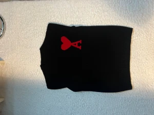 Svart stickad tröja med rött hjärta - Svart stickad tröja från AMI Paris med ett stort rött hjärta och A på bröstet. Tröjan har ribbade muddar vid hals, ärmslut och nederkant. Klassisk passform och mjukt material som känns skönt mot huden. Perfekt för dig som gillar stilrena detaljer. Storlek M 