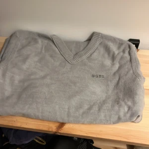 Grå v-ringad ull tröja från BOSS - Snygg grå v-ringad ull tröja från BOSS med diskret broderad logga på bröstet. Tröjan är stickad i mjuk ull och har ribbade muddar vid hals, ärmslut och nederkant. Perfekt för dig som gillar stilren och klassisk look..