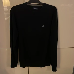 Svart stickad tröja från GANT - Svart stickad tröja från GANT i storlek L. Klassisk rund hals och diskret logga broderad på bröstet. Tröjan har långa ärmar och ribbade muddar vid hals, ärmslut och nederkant. Perfekt för dig som gillar stilrena och enkla plagg.