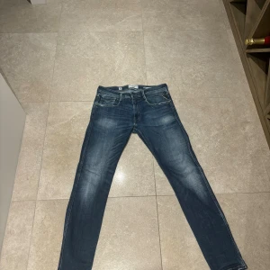Replay Anbass blå jeans slim fit - Snygga blå Replay Anbass jeans med slim fit och klassisk femficksdesign. Jeansen har en mörkblå tvätt med lätt slitna detaljer och kontrastsömmar. Skinnpatch med Replay-logga baktill i midjan. Perfekta för dig som gillar stilrena och moderna jeans.