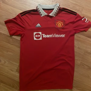 Manchester United röd fotbollströja Adidas - Säljer en officiell Manchester United fotbollströja från Adidas i rött med vit krage och svarta detaljer på axlarna. Tröjan har klubbmärke, TeamViewer-tryck och Adidas-logga på bröstet. Materialet är lätt polyester, perfekt för match eller träning.