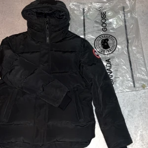 Svart dunjacka från Canada Goose M - Säljer en svart dunjacka från Canada Goose i storlek M. Jackan har huva, dragkedja och ribbade muddar vid ärmslut. Klassisk patch på ärmen och flera fickor framtill. Perfekt för kalla dagar och har en clean, stilren look.