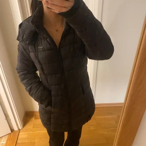 Svart lång pufferjacka från Marikoo - Svart lång pufferjacka från Marikoo. Camo-mönstrat foder i rött, grönt, beige och svart. Jackan har stor luva, långa armar, 2 dragkedjor framtill som går att knäppa upp i längden. Basic men snygg quiltad design och praktiska fickor på sidorna. Perfekt för långa kalla dagar ute i kylan. Jackan är använd få gånger och kommer med extra knappar. Pris kan diskuteras (Ordinarie pris 1150)😇