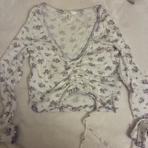 Blommig topp - Supersöt topp från H&M i storlek XS, med blommigt mönster i lila och grönt på vit bakgrund. Toppen har långärmade ärmar, v-ringning och snörning framtill som ger en croppad look. Man kan även ändra på snöret så att tröjan kan blir antingen längre eller kortare. Perfekt för vår och sommar! Pris kan diskuteras!