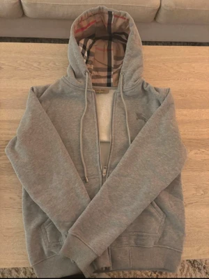 Grå Burberry hoodie med dragkedja - Snygg grå hoodie från Burberry med klassiskt rutigt foder i huvan och insidan. Dragkedja framtill, broderad Burberry-logga på bröstet och två fickor. Mjuk och skön bomullsblandning, perfekt för chill dagar. Storlek medium.