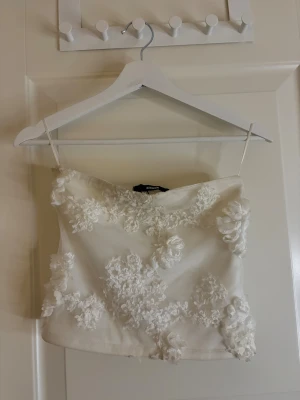 Vit blommig tubtopp från Bikbok - Supersöt vit tubtopp från Bikbok i storlek XS,med blommor och broderade detaljer. Finns dragkedja med. Köpt i somras och andvänt ett par gånger men det finns inga hål eller trasigheter