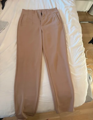 Beige kostymbyxor Streetwear Society XS - Snygga beige kostymbyxor från Streetwear Society i storlek XS. Byxorna har hög midja, raka ben och pressveck framtill. De är tillverkade i ett mjukt material med två fickor på sidorna och stängs med knapp och dragkedja.