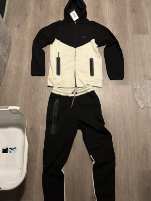Nike svart och vit tracksuit  - Nike tracksuit i svart och vit med bred resår i midjan och dragkedja på ena benet. Snygg kontrastdesign med beige detaljer på vaderna och midjan. Perfekta för chill eller träning, tillverkade i mjukt bomullsmaterial.