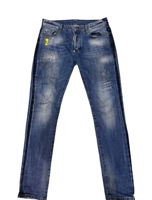 Dsquared2 Jeans - Snygga blå jeans med slitningar, färgstänk och ett gult tryck med siffran 2 framtill. På bakfickan finns broderade detaljer med flagga, glas och burk. Jeansen har en rak passform och klassisk femficksdesign. Perfekt för dig som gillar unika detaljer och streetstyle.