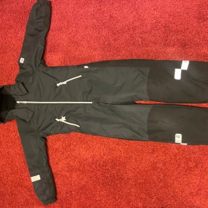 Reima-TEC Finn Kiddo svart vinteroverall, storlek 140 - Svart Reima vinteroverall med dragkedja framtill och två fickor med dragkedja. Modellen heter Reima-TEC Finn Kiddo.   Reflexer finns på ärmar, ben, rygg och på blixtlåsen fram. Luvan är avtagbar.   Förstärkta partier på bakdel, ben och underarmar.   Inbyggda hängslen. Perfekt att ta av ovandelen när man är i värmestugan och ha hängslena på.   Reima är ett mycket bra märke med en väldigt bra passform. Finsk mycket hög kvalitet.   Vind- och vattentät.   Använd ett fåtal gånger  Nypris ca 1600 kr. 