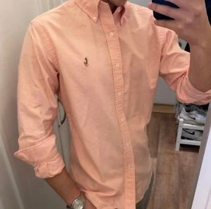 Rosa Skjorta från Polo Ralph Lauren (Slim Fit) - Snygg rosa skjorta från Polo Ralph Lauren i slim fit. Klassisk button-down krage, broderad logga på bröstet och vita knappar framtill. Skjortan är långärmad och tillverkad i mjuk bomull som känns skön mot huden. Perfekt för dig som gillar stilrena plagg med lite färg. Storlek M Snabb Leverans!