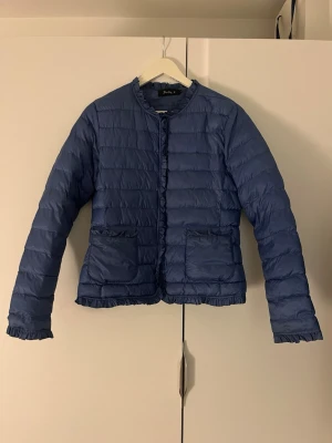 Blå dunjacka från Softy - Dunjacka i mörkblått från Softy med volangdetaljer vid krage, fickor, ärmslut och nederkant. Jackan har två stora fickor framtill och stängs med dragkedja. Det står att den är i storlek L men jag skulle absolut säga att den är M