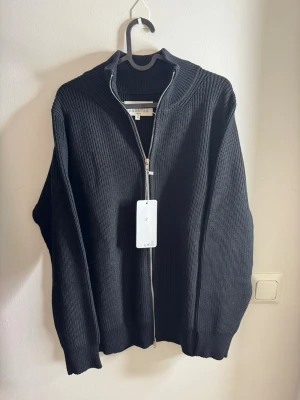 Svart stickad kofta från Maison Margiela med zip up - Riktigt snygg svart stickad kofta från Maison Margiela med ribbad struktur och silverfärgad dragkedja hela vägen. Klassisk ståkrage och de ikoniska vita sömmarna i nacken. Perfekt till lager på lager och ger en clean look. Size 44 (s)