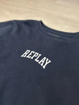 Mörkblå sweatshirt från Replay storlek M - Snygg mörkblå sweatshirt från Replay med vit logotyp tryckt på bröstet. Tröjan har rund hals, långa ärmar och ribbade muddar vid ärmslut och nederkant. Tillverkad i mjuk bomull och polyesterblandning som känns skön mot huden.