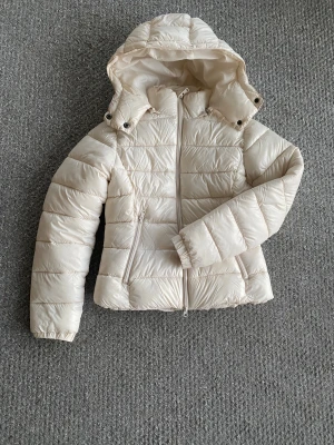 Beige pufferjacka med huva - Säljer en beige pufferjacka med huva och dragkedja framtill. Jackan har två sidofickor med dragkedja och är quiltad för extra värme. Perfekt för kyliga dagar och har en stilren look som passar till det mesta.