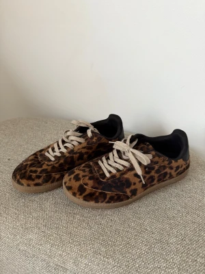 Leopardmönstrade sneakers från H&M - Snygga sneakers från H&M med leopardmönster i bruna och svarta toner. Skorna har vit snörning, rund tå och platt sula. Ovandelen är i mockaliknande material och insidan är svart. 