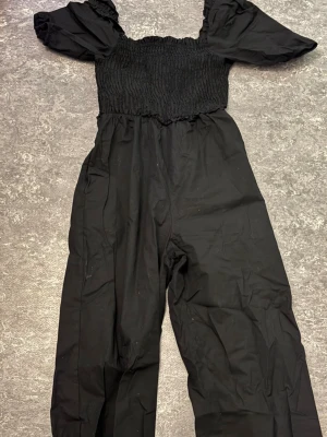 Svart byxdress med puffärm - Snygg svart byxdress med puffärmar och smockad överdel. Byxorna är raka och dressen har en cool, enkel look som passar perfekt för dig som gillar stilrena plagg. Materialet känns lätt och bekvämt mot huden.