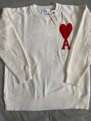 Vit stickad tröja från Ami Paris - Vit stickad tröja från Ami Paris med rund halsringning och ribbade muddar. På bröstet finns ett stort rött hjärta med bokstaven A. Tröjan har en clean look och är perfekt för dig som gillar stilrena detaljer.
