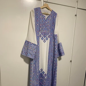 Vit långklänning med blått broderi - En palestinisk vit långklänning med detaljerat blått broderi och inslag av rött och gult. Klänningen har vida långärmar och ett geometriskt mönster längs hela plagget. Perfekt för dig som gillar traditionell design med modern twist. (Den har kommit till användning en kväll under 2 timmar). Köpte för några tusen och de drf lägsta priset kommer vara 1000kr.