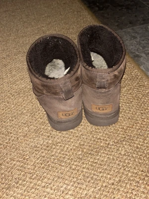 Bruna UGG boots i mocka - Mysiga bruna UGG boots med rund tå och platt sula. Utsidan är i mjuk mocka och insidan är fodrad med mörkt fluffigt material som håller fötterna varma. 